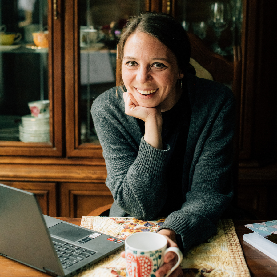 A woman, Robyn Roste, smiling at the camera, leaning on a table with a laptop, papers, and a mug in a cozy, well-lit room.