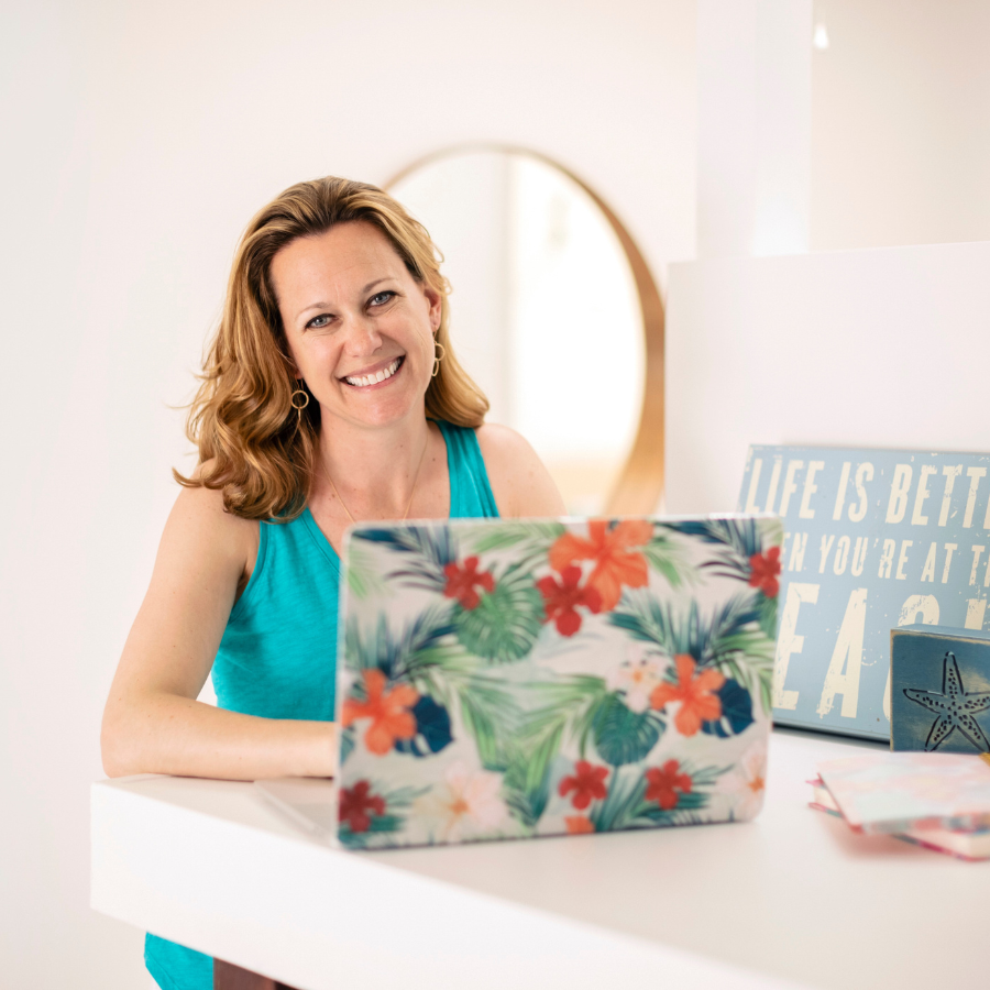 A woman in a turquoise tank top is smiling while working on a laptop with a colorful floral cover. On the desk, alongside a book and a sign that reads "LIFE IS BETTER WHEN YOU'RE AT THE BEACH," she has her February 2024 Income Report open, displaying her latest financial achievements.