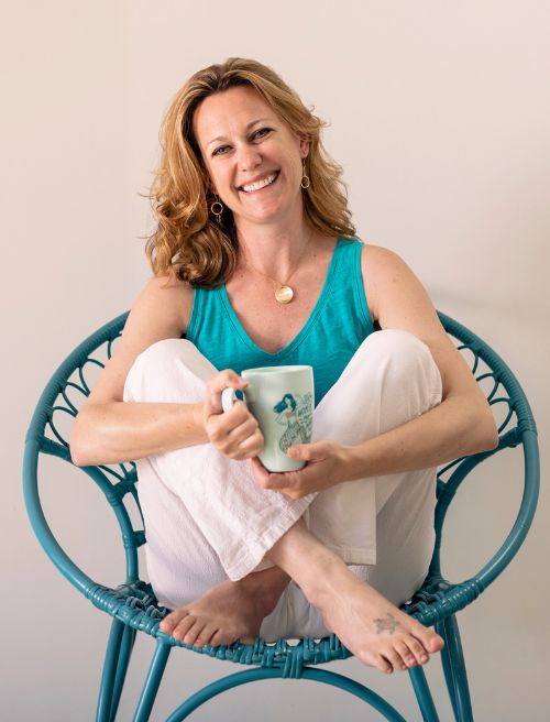 A woman with light brown hair sits on a blue chair, smiling, wearing a turquoise tank top and white pants, holding a white mug with a design, and appearing relaxed.