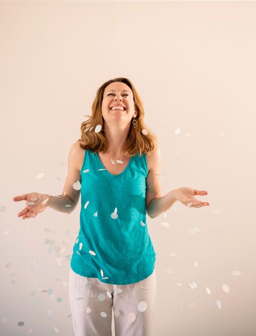 A woman in a teal sleeveless top and white pants stands smiling as paper confetti falls around her.