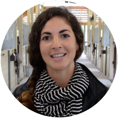 alissa mayer headshot A woman with long curly hair in a braid, wearing a black and white striped scarf and a dark jacket, stands in a horse stable aisle with stalls in the background.