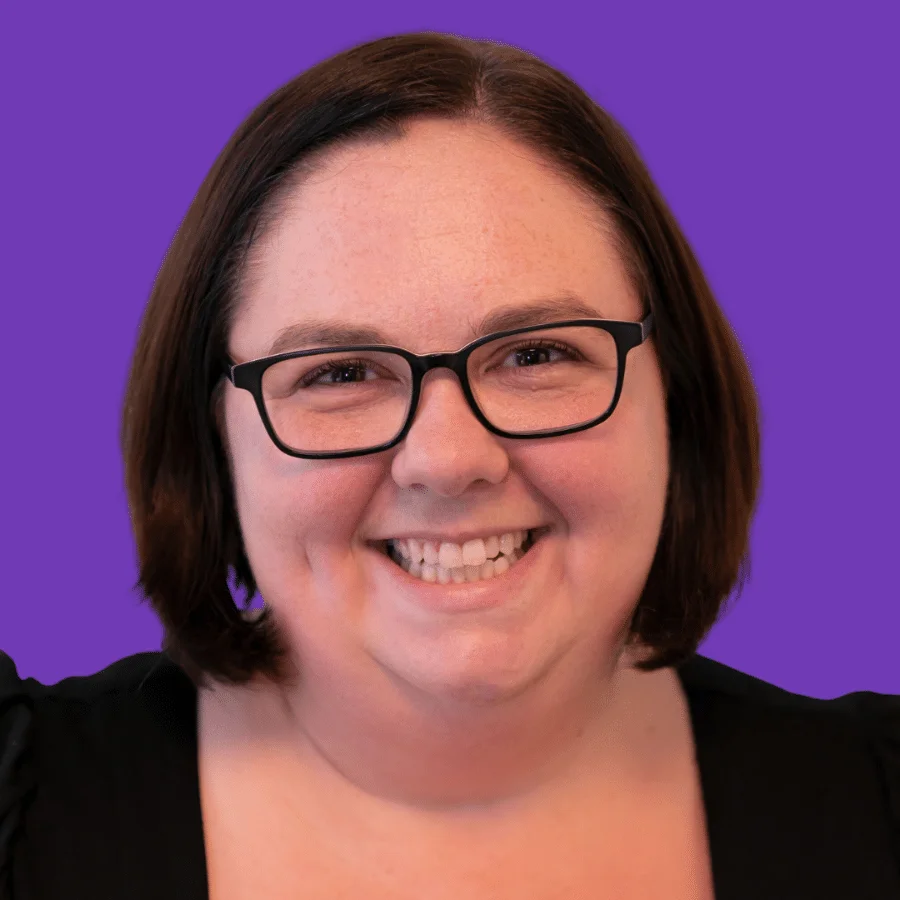 Adrienne Farrow, a woman with shoulder-length brown hair, wearing glasses and a black top, smiles at the camera against a purple background.