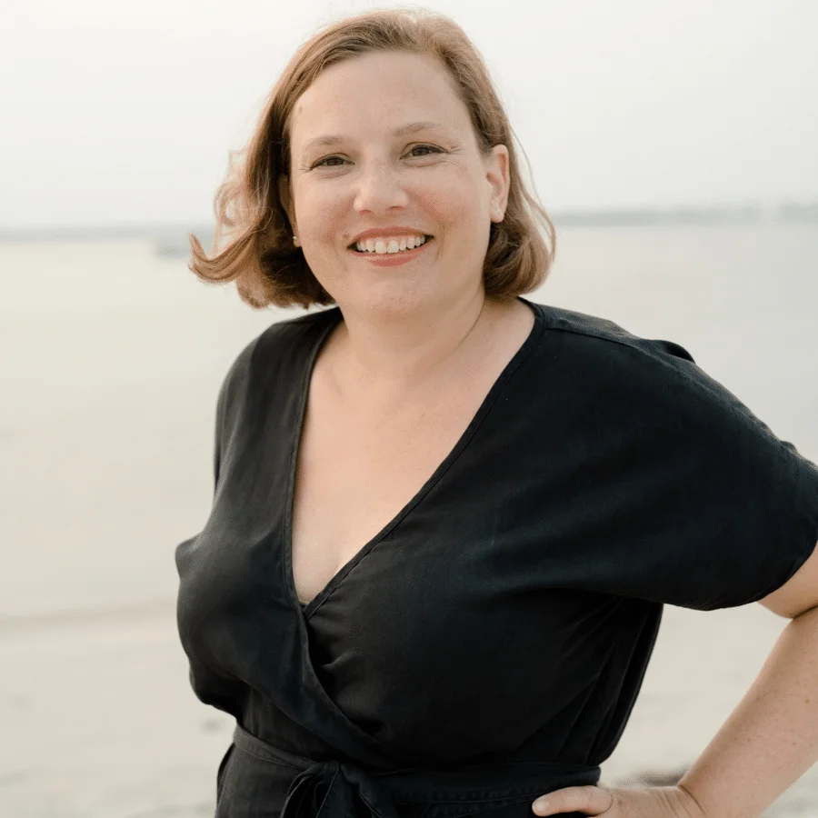 Kate Gilbert, a woman with short, light brown hair, is standing on a beach, smiling and looking at the camera. She is wearing a black wrap dress. The background shows a pale sky and calm water.