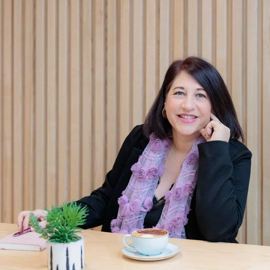 Sabine Matharu, a woman with shoulder-length dark hair, wearing a purple scarf and black top, sits at a table with a cappuccino, a potted plant, and a closed book. She is smiling and facing the camera.